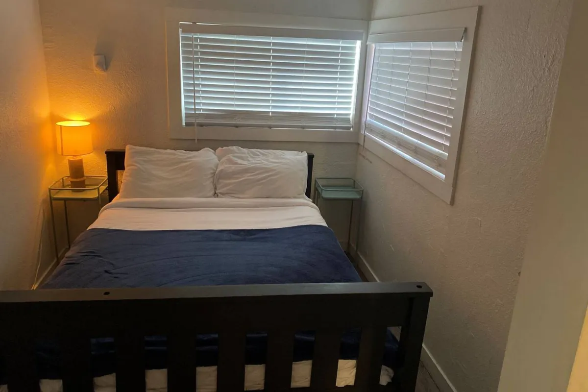 A bright, tranquil bedroom with a plush bed dressed in neutral linens, flanked by a modern nightstand and a softly glowing lamp. Natural light pours in, creating a soothing ambiance perfect for winding down after a day at the beach.