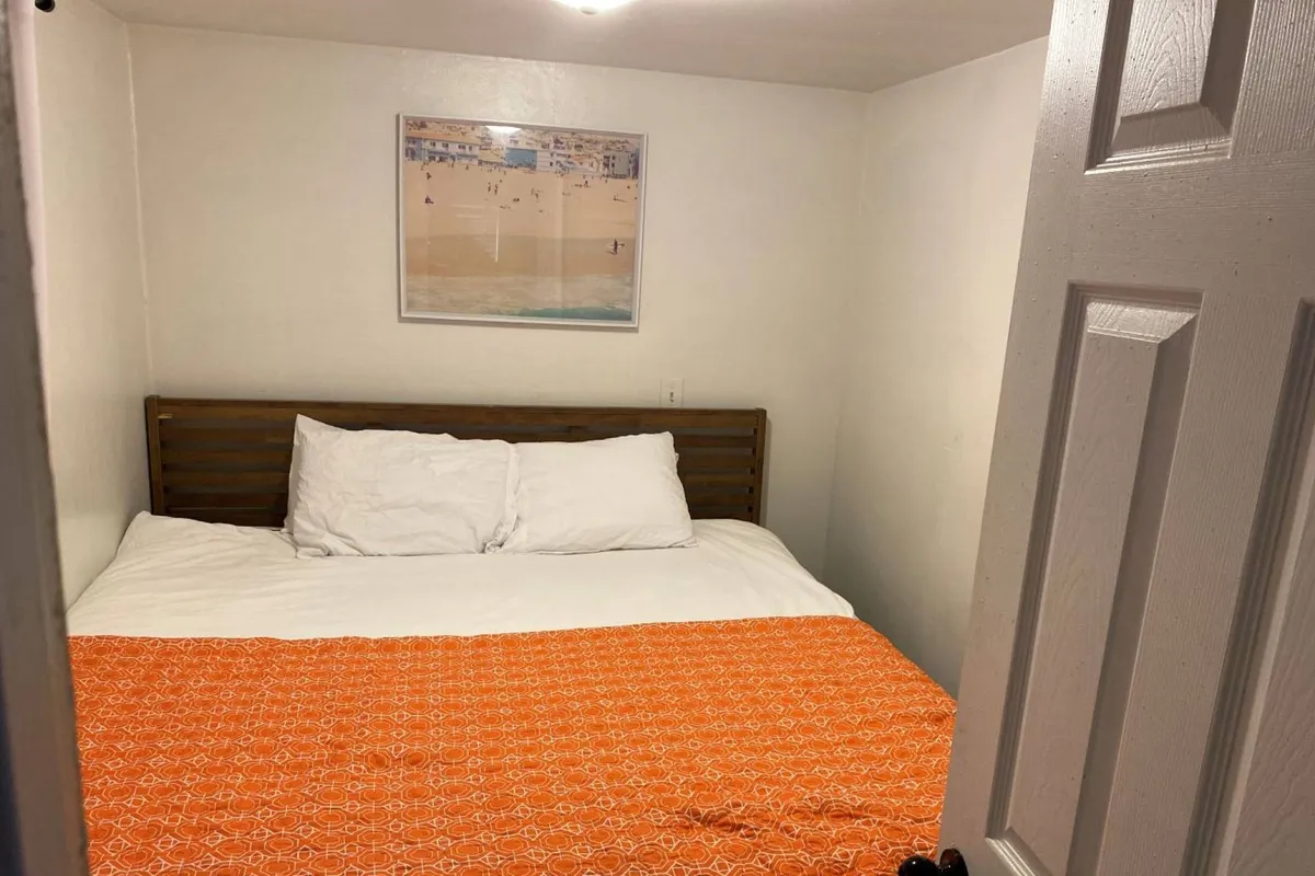 A cozy bedroom featuring a full-sized bed adorned with a vibrant orange blanket and a framed beach-themed picture above the headboard. Sunlight streams into the inviting space, suggesting a cheerful spot for both rest and relaxation.