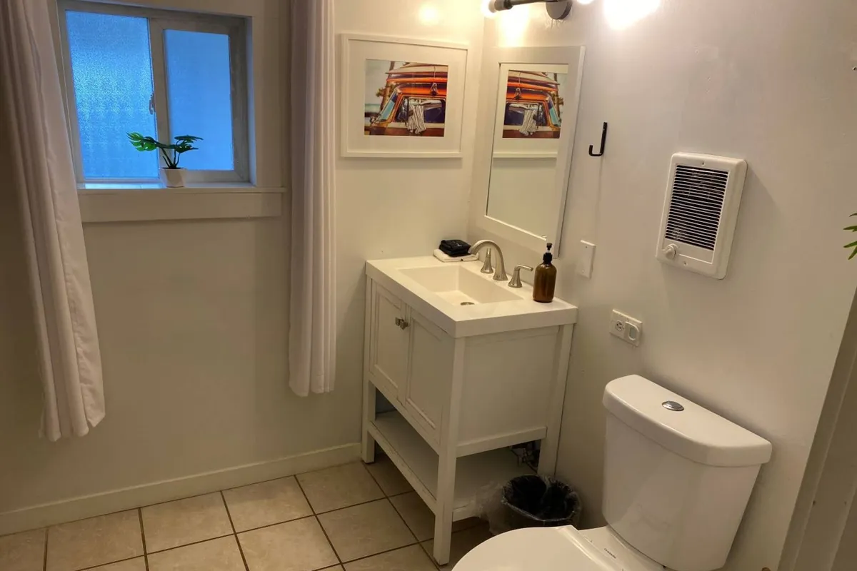 A fresh, inviting bathroom featuring a vanity with a sink, framed artwork, and a window with a leafy plant. The space is bright and airy, setting the scene for refreshing morning rituals or unwinding in the evening.