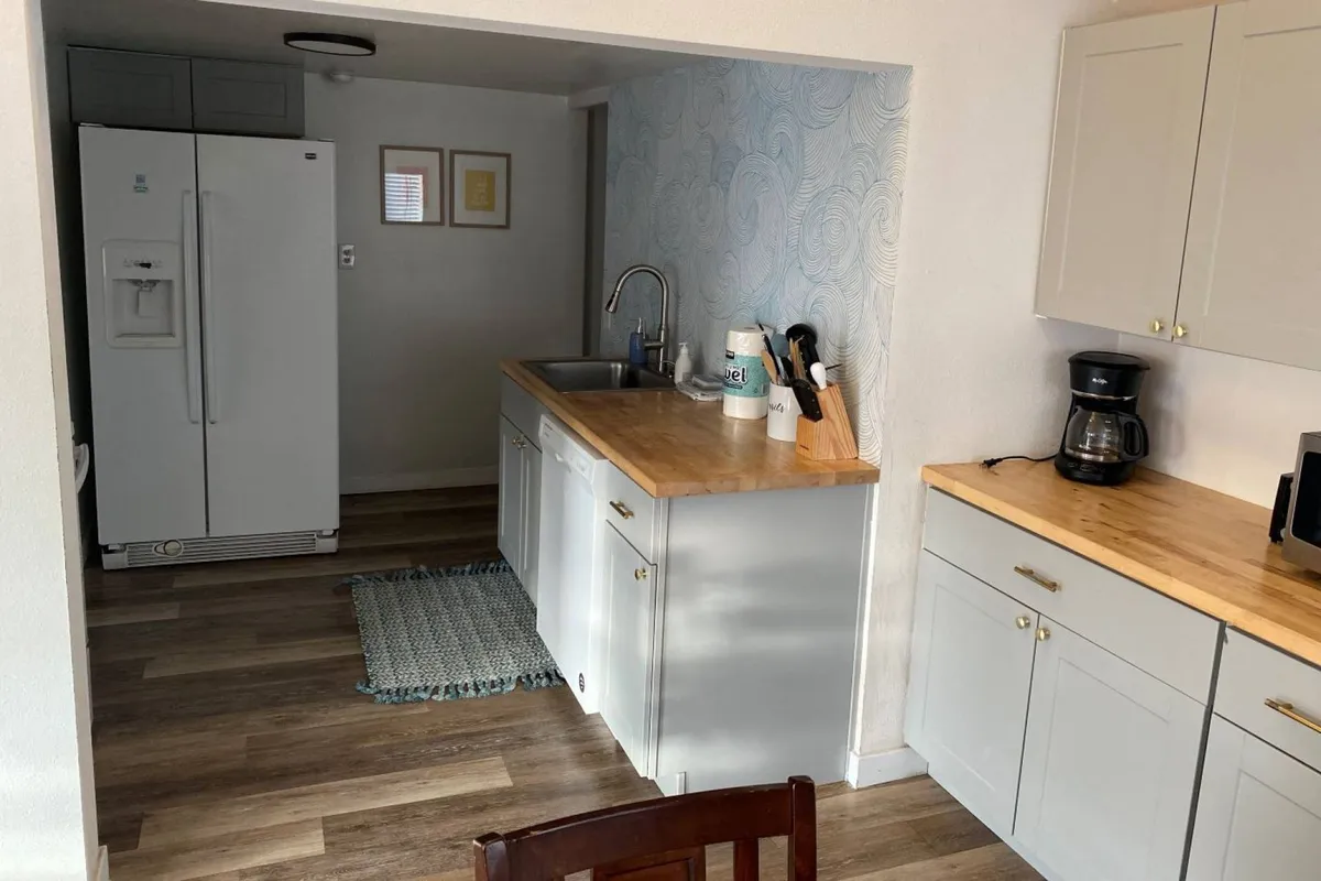 This image showcases a cozy, functional kitchen with a visible sink, refrigerator, countertop, and coffee maker. The bright, inviting space is well-suited for preparing meals or enjoying a morning coffee, embodying the idea of a well-equipped kitchen within an open-concept layout. The arrangement encourages socializing and shared activities, aligning with the description of the heart of the home.